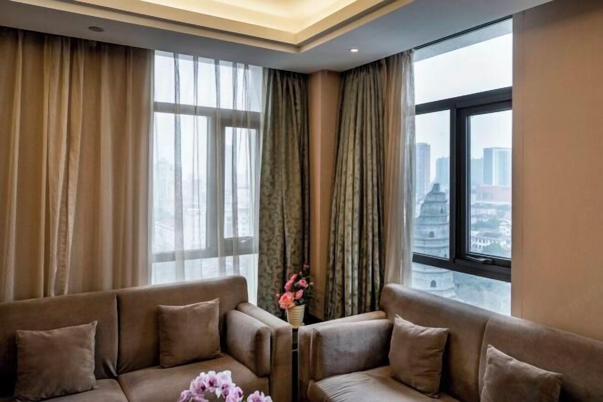 A modern living room with a sofa set, a vase with flowers, and a city view through large windows.