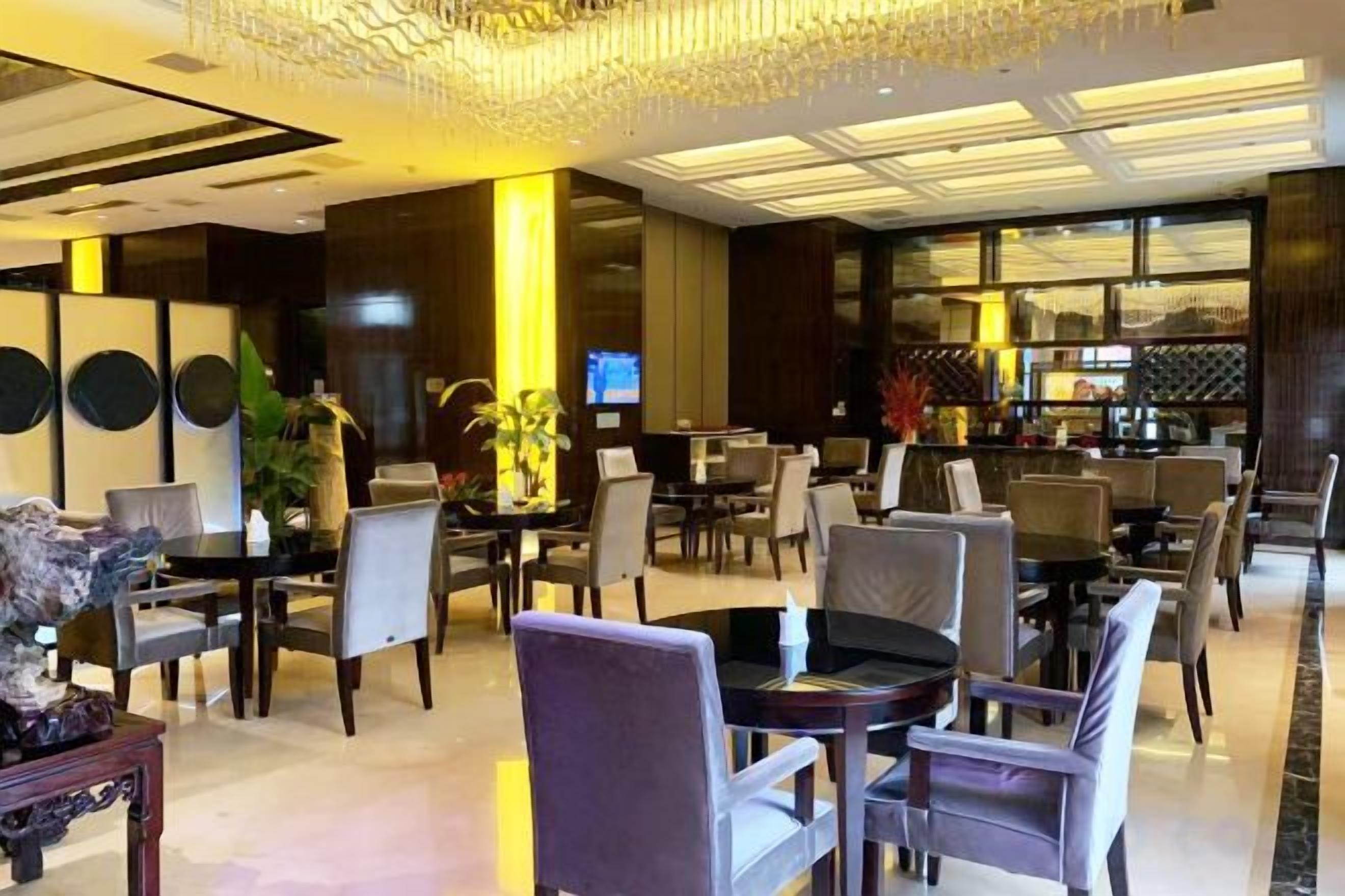 A modern hotel lobby with round tables, comfortable seating, a bar area, and a television screen.