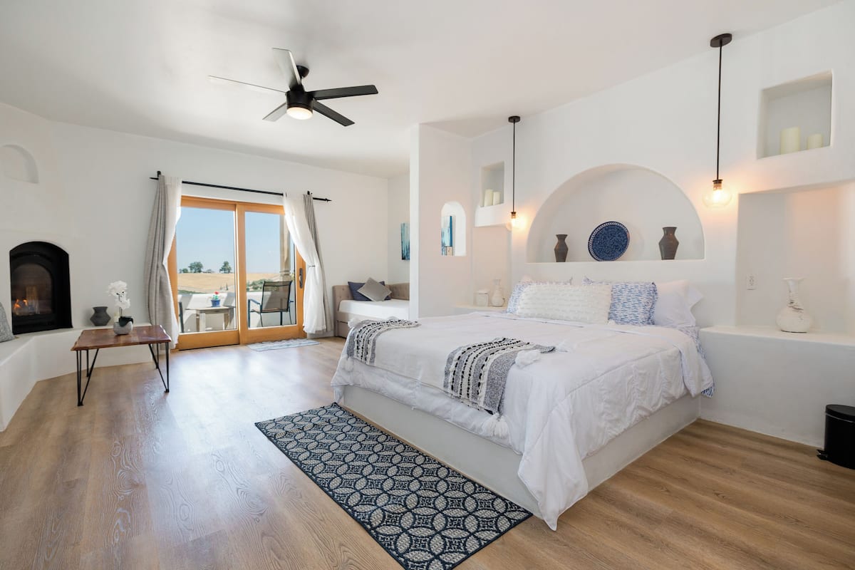 A bedroom with a bed, a ceiling fan, a fireplace, and a view through a glass door.