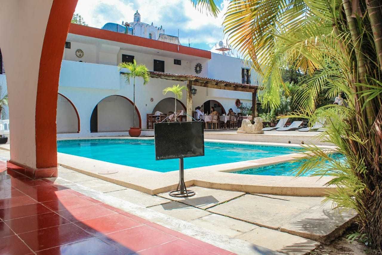 Una piscina con zona de estar cubierta, palmeras y un edificio con arcos al fondo.