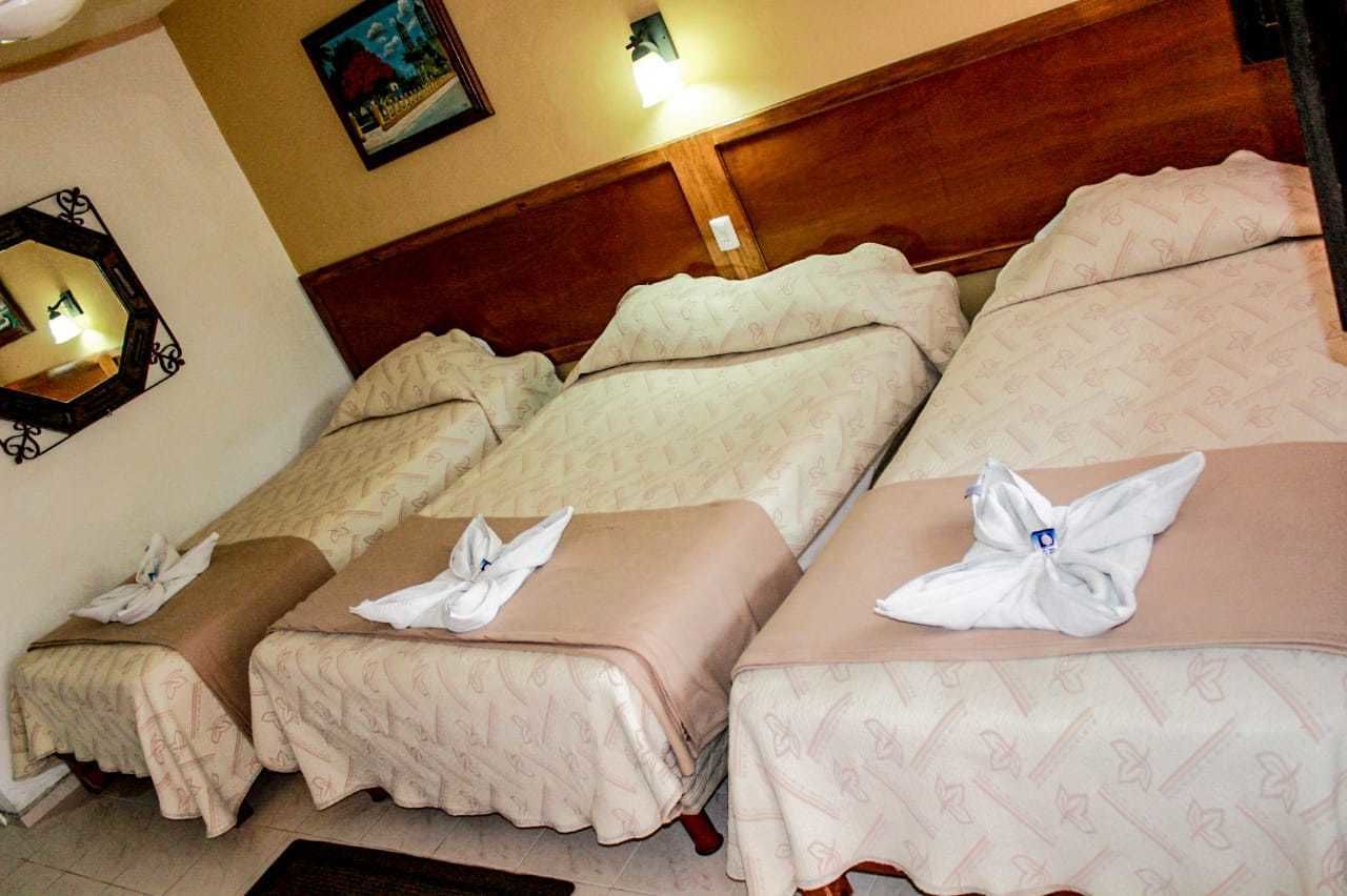 Ein Hotelzimmer mit drei Betten, jeweils mit beigefarbener Bettwäsche und weißen Handtüchern, die obenauf gefaltet sind.