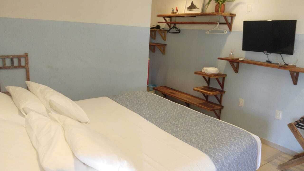 Ein Schlafzimmer mit einem Bett, Holzbanken, einem an der Wand montierten Fernseher und Regalen mit diversen Gegenständen.