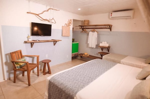 A hotel room with a bed, a chair, a small table, a TV, and a shelf with towels and a basket.