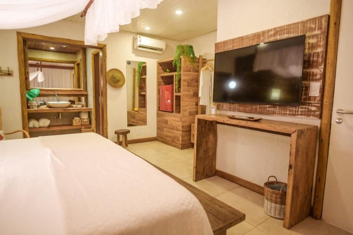 Une chambre à coucher avec un lit, un téléviseur posé sur un meuble en bois, un miroir, une étagère avec des articles de toilette et un petit tabouret.
