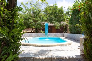 Piscina com fonte, cercada por vegetação exuberante e um deck revestido de azulejos.