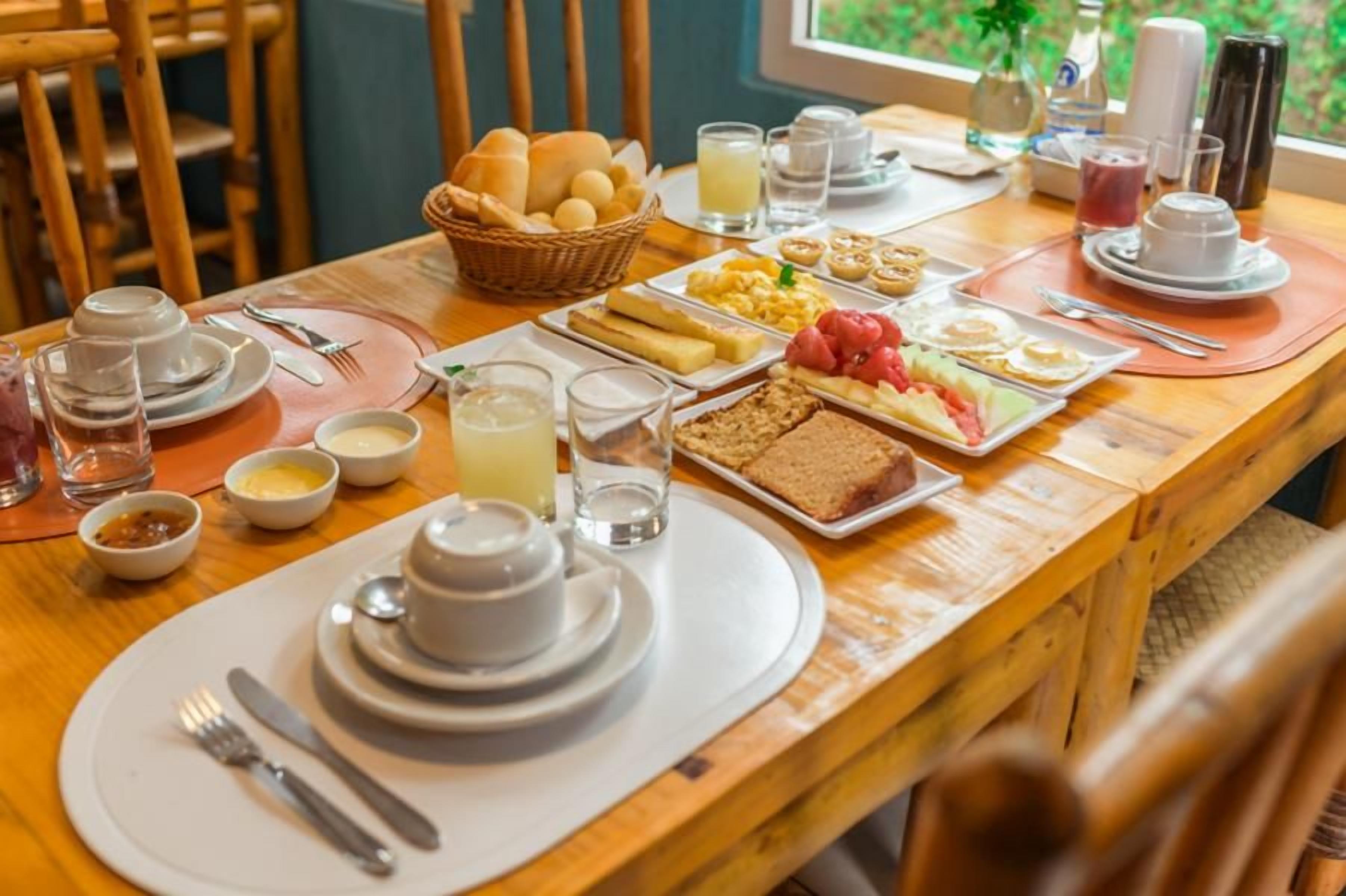 A breakfast setting with a variety of food items, including bread, eggs, and fruits, arranged on a wooden table with chairs around.