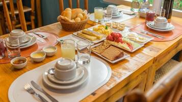 A breakfast setting with a variety of food items, including bread, eggs, and fruits, arranged on a wooden table with chairs around.