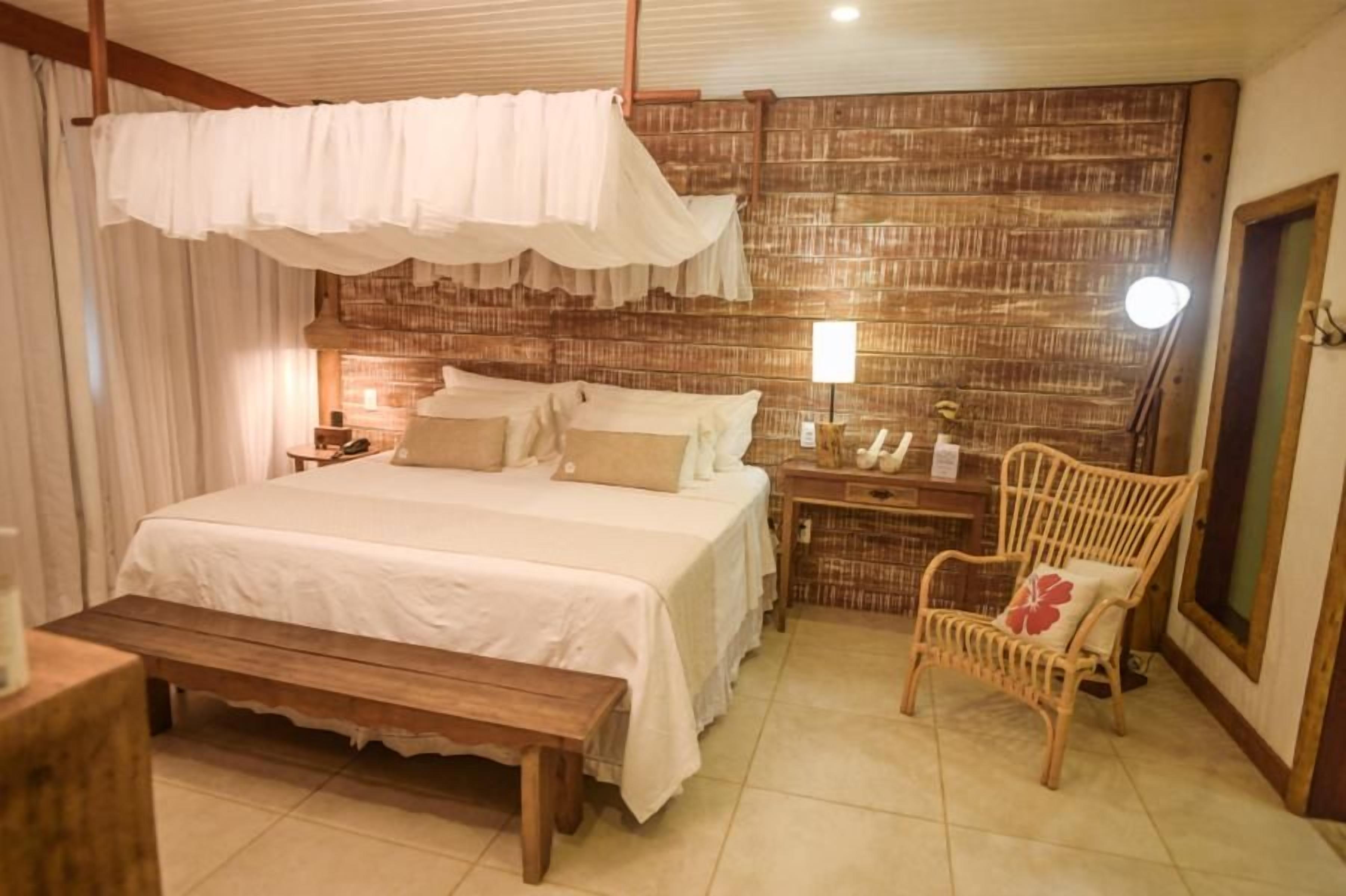 A bedroom with a large bed, a wooden bench, a wicker chair, and a nightstand with a lamp.