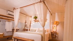 A cozy bedroom with a canopy bed, a bedside table, a hanging plant, and a hat on the wall. - Ecocharme Pousada Do Marcilio (Fernando de Noronha)