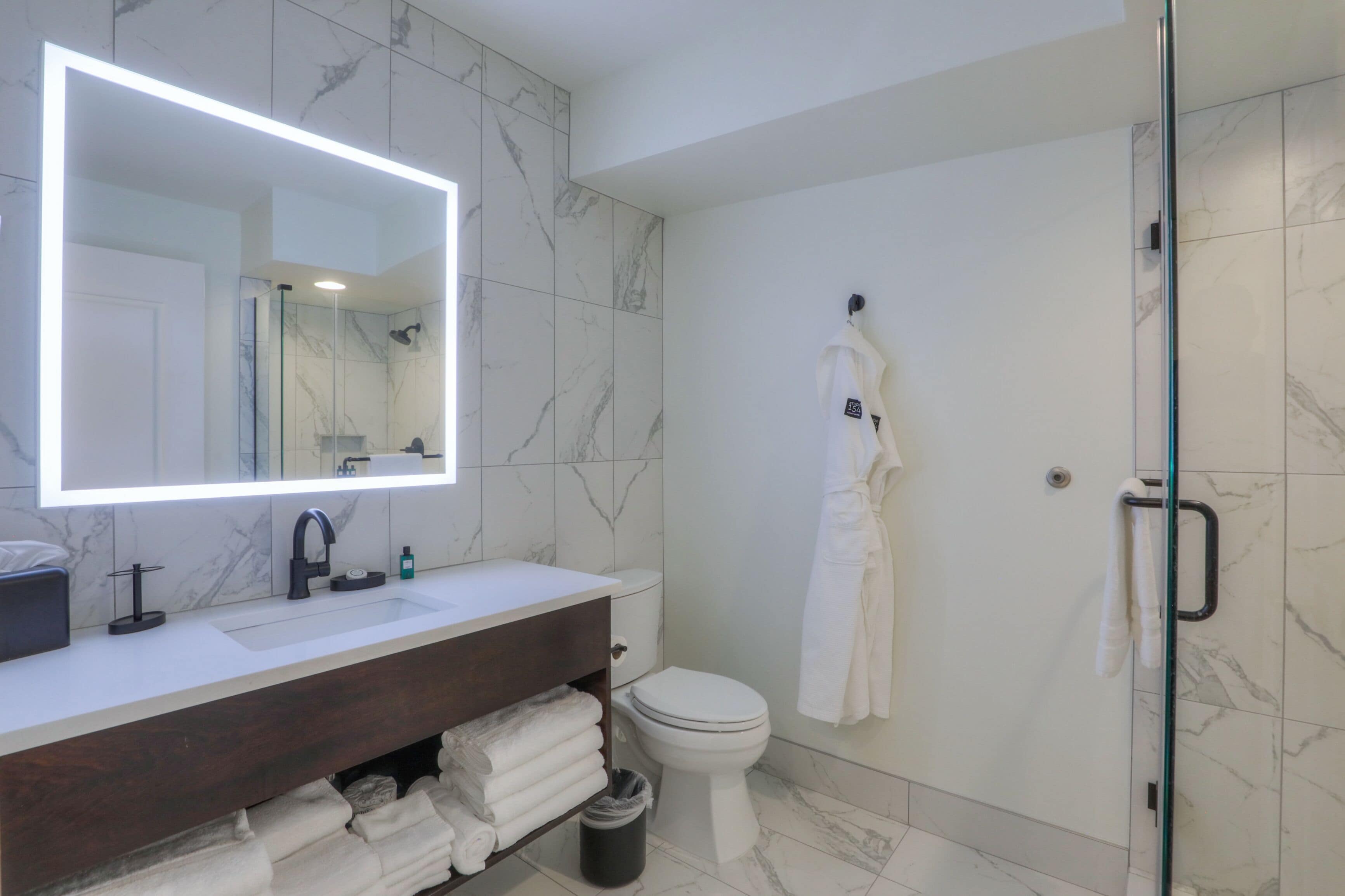 A modern bathroom with a marble-tiled shower, a white sink with a dark vanity, and a large mirror with integrated lighting.