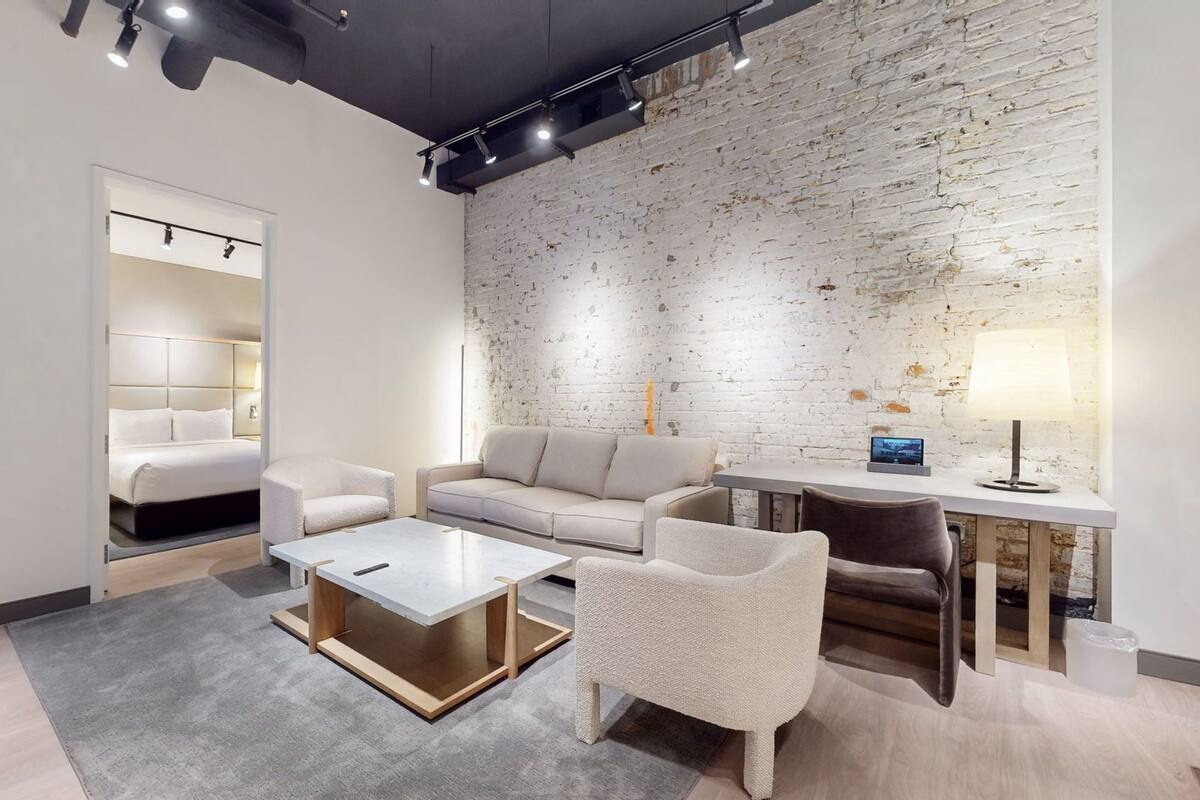 A modern hotel room with a white brick wall, a sofa, a coffee table, and a desk with a lamp.