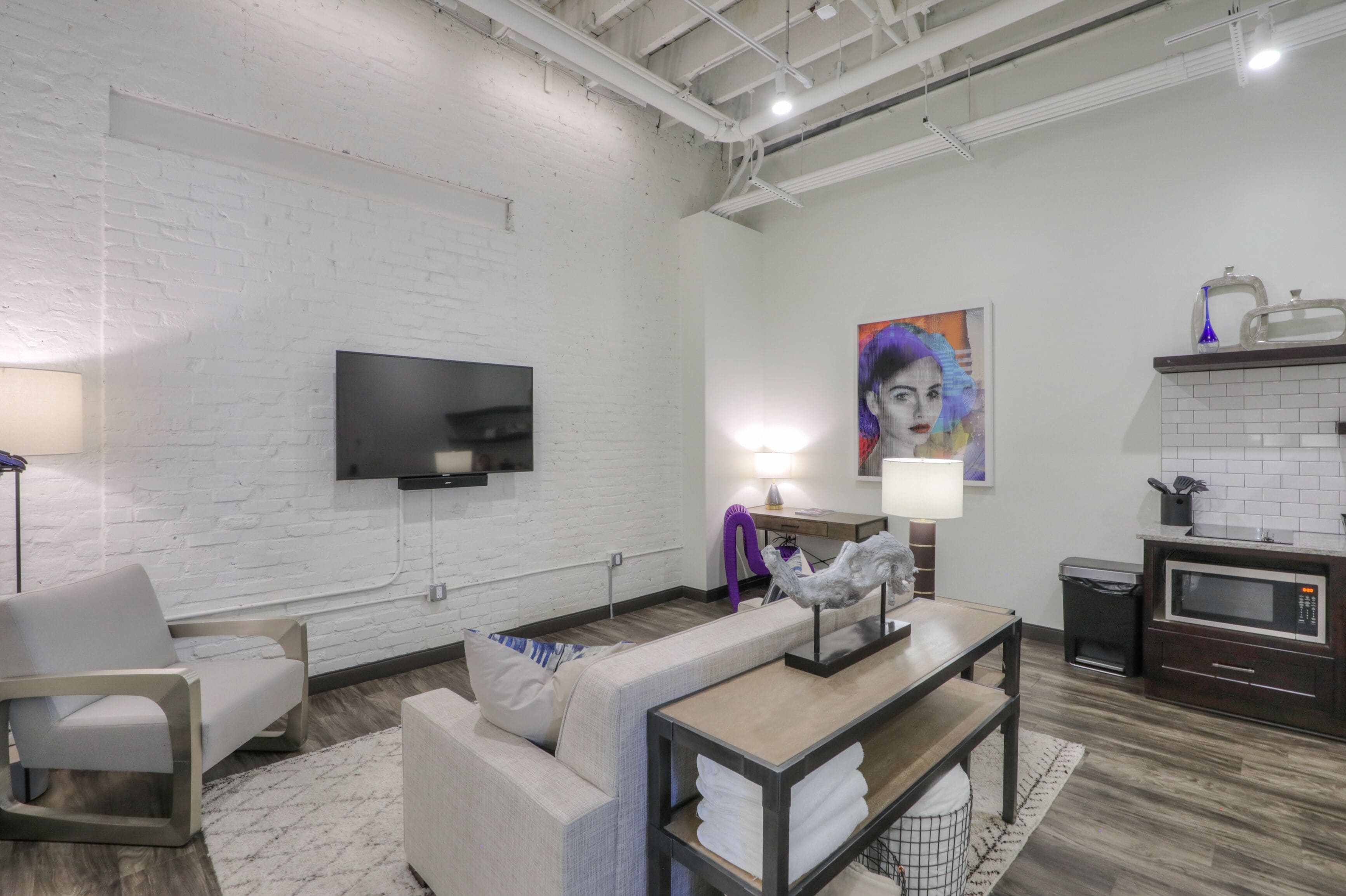 A modern living room with a white brick wall, a flat-screen TV, a sofa, a coffee table, and a kitchen area with a microwave.