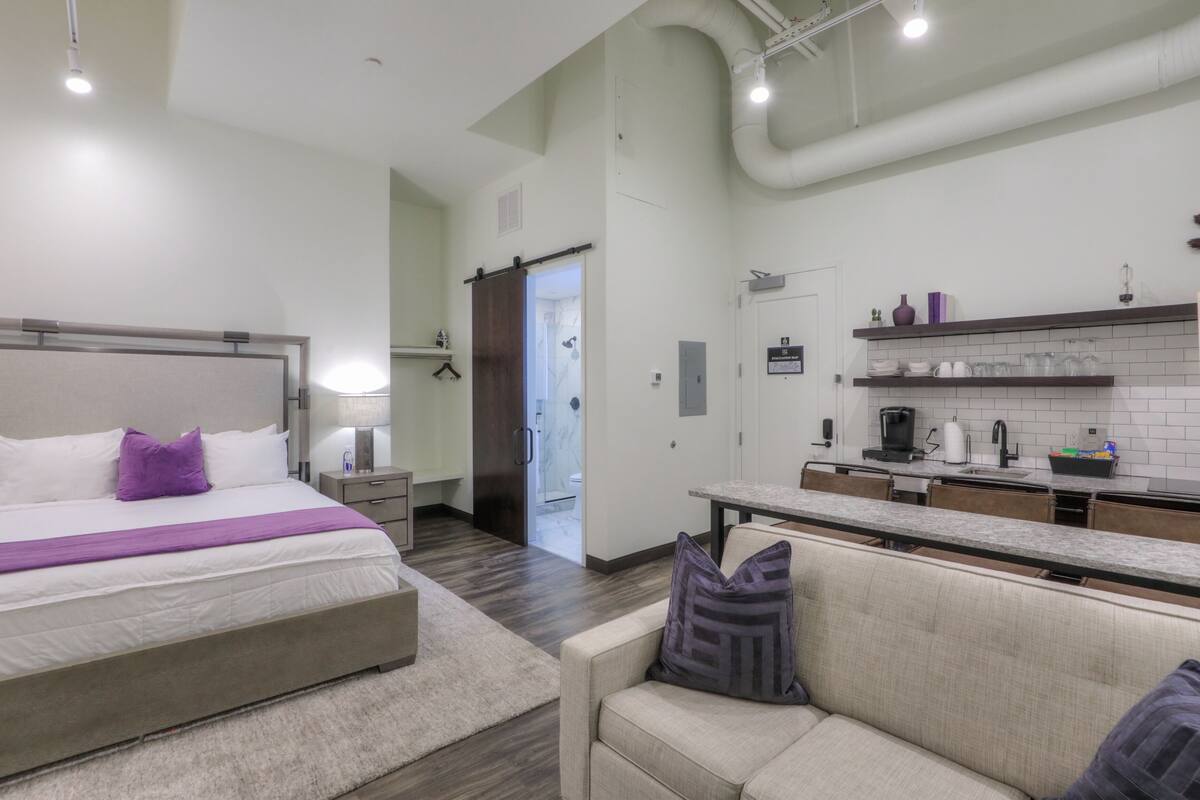 A modern hotel room with a large bed, a sofa, a kitchenette, and a bathroom visible through an open door.