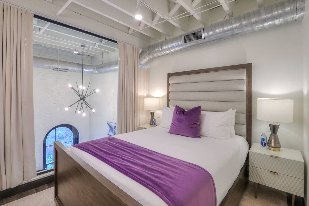 A modern hotel room with a large bed, a view through a glass wall, and a ceiling with exposed ductwork.