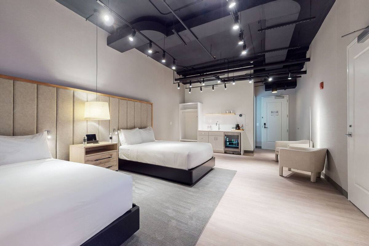 A modern hotel room with two beds, a desk, a chair, and a kitchenette.