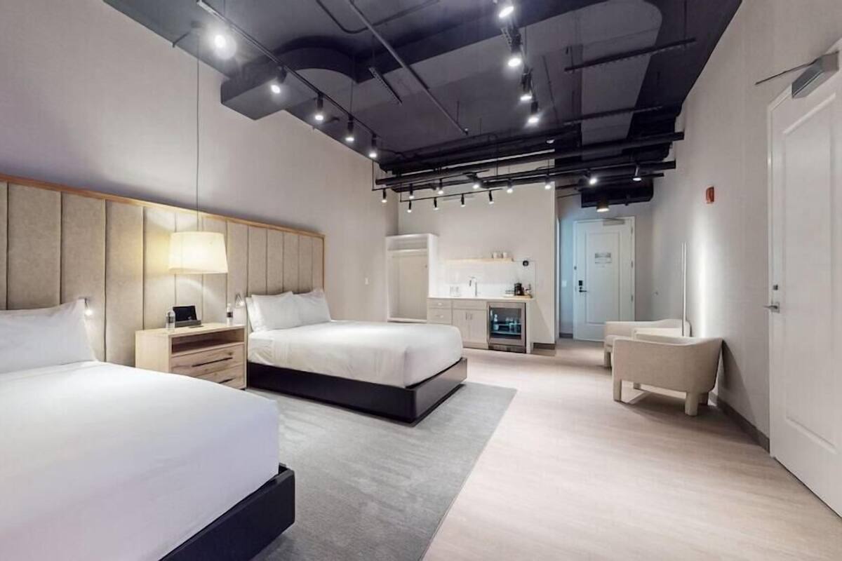 A modern hotel room with two beds, a desk, a chair, and a kitchenette.