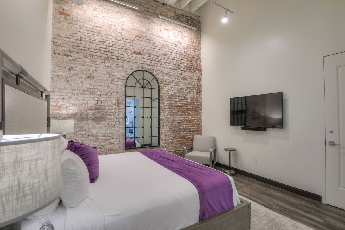 A modern hotel room with a brick wall, a large arched window, a bed with white linens and a purple throw, a white armchair, a small round table, and a flat-screen TV mounted on the wall.