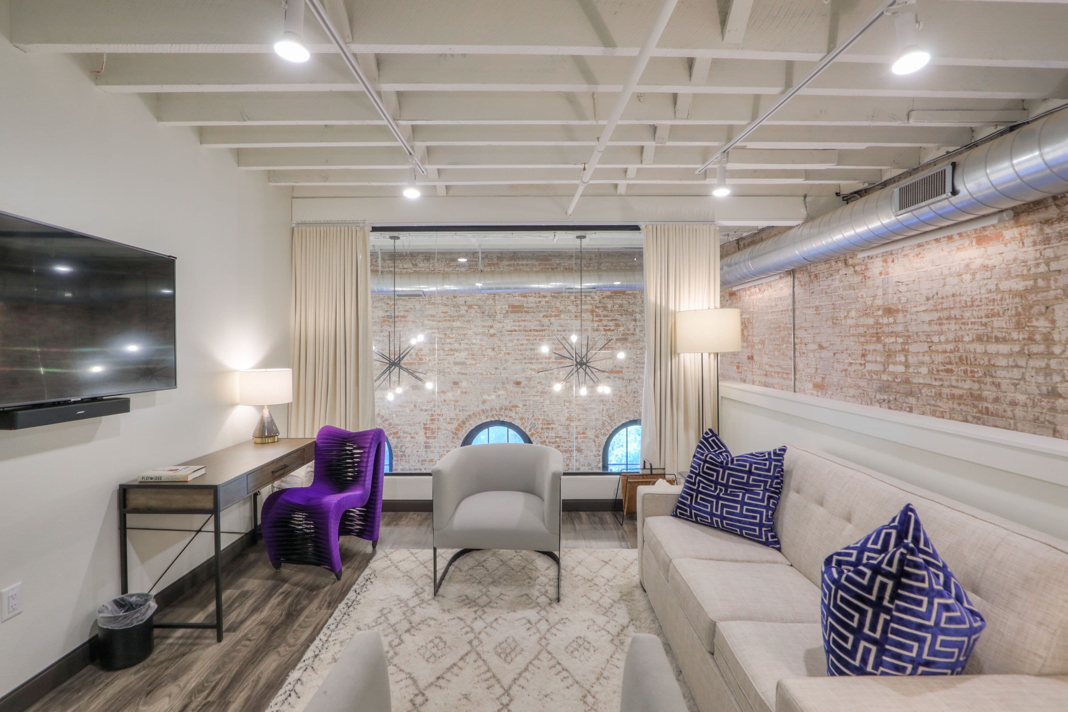 A modern living room with a brick wall, a flat-screen TV, a sofa, a chair, and a desk.