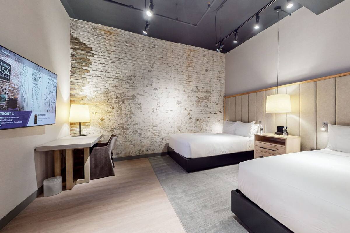 A modern hotel room with a brick accent wall, a large bed, a smaller bed, a desk with a chair, and a nightstand with a lamp.