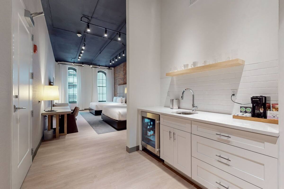 A modern hotel room with a kitchen area, a dining table, and a large bed.