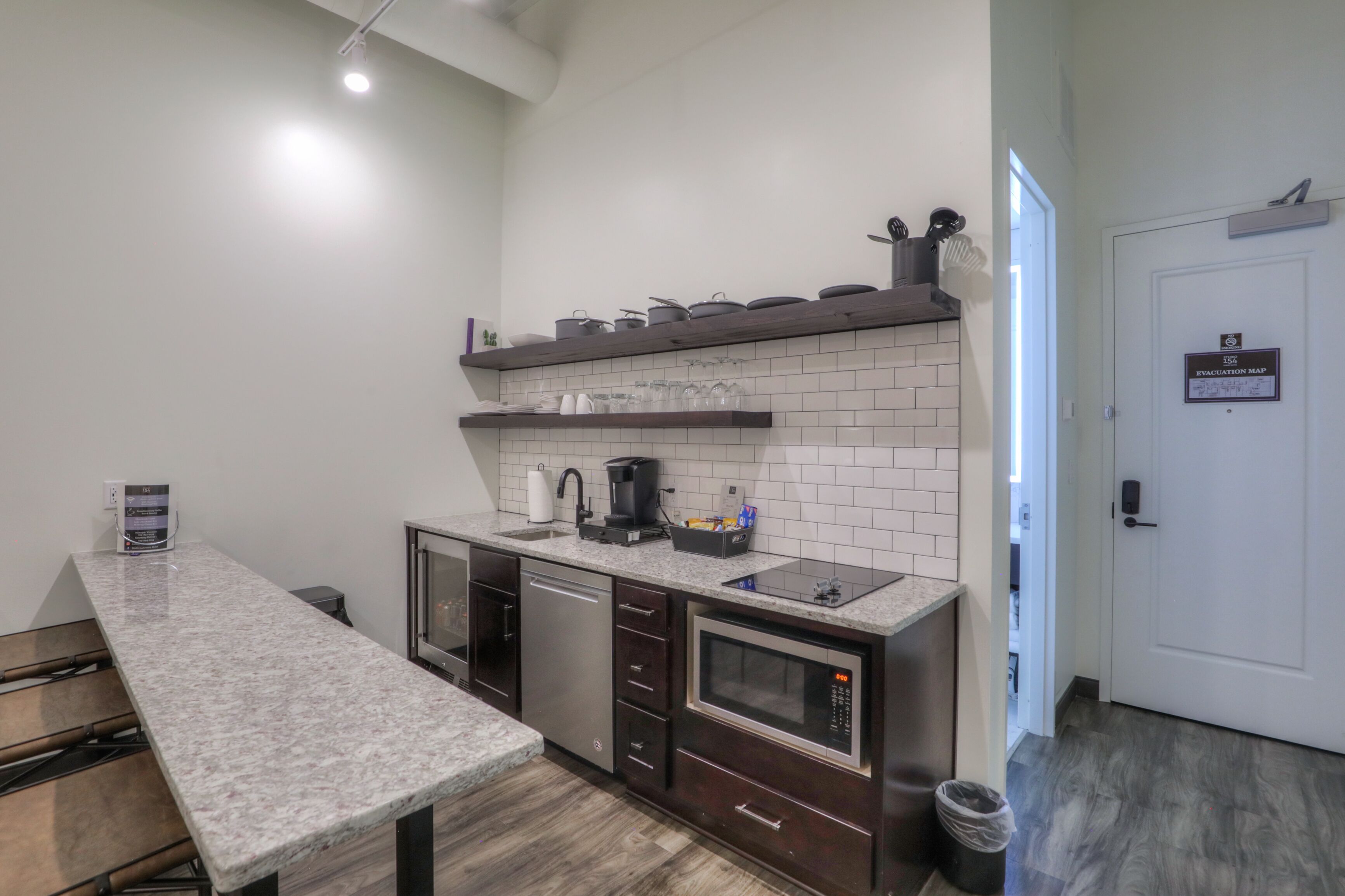 A modern kitchen with a granite countertop, built-in dishwasher, microwave, and a coffee maker. There is a shelf above the sink with kitchenware and a door with an evacuation map sign.