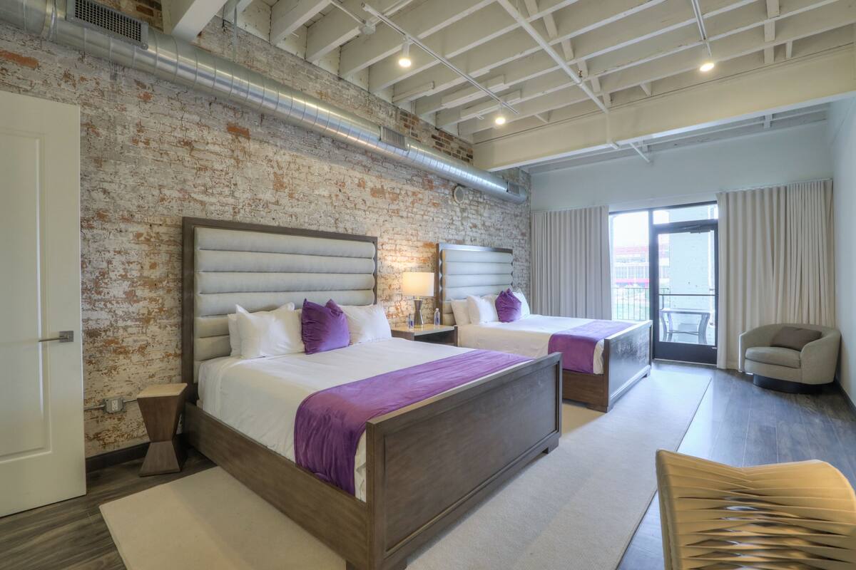 A modern hotel room with a brick wall, two beds with headboards, a wooden floor, a chair, and a large window with curtains.