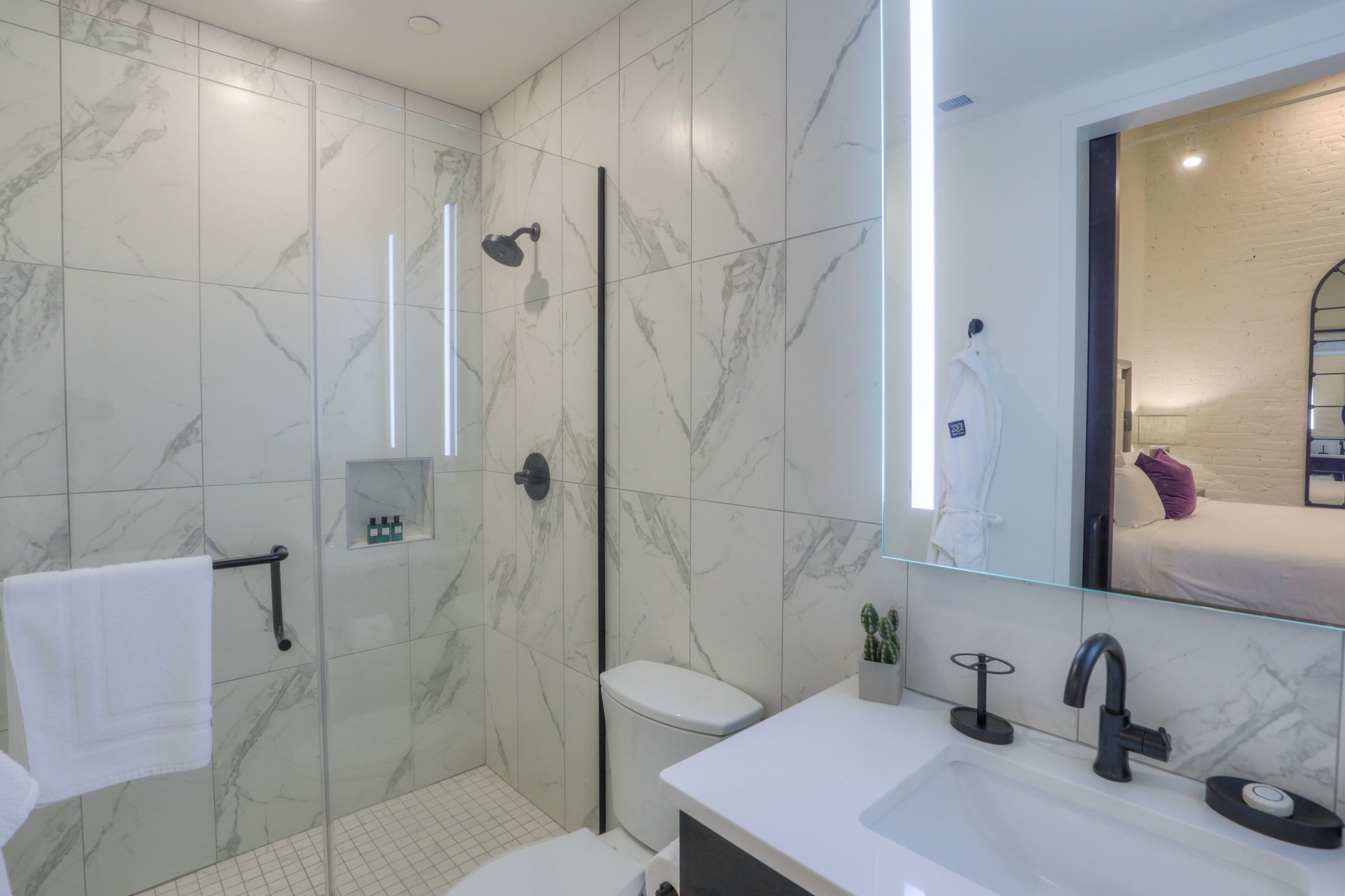 Un baño moderno con ducha de vidrio, un lavamanos blanco y un espejo grande que refleja una cama con un cojín morado.