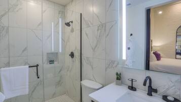Un baño moderno con ducha de vidrio, un lavamanos blanco y un espejo grande que refleja una cama con un cojín morado.