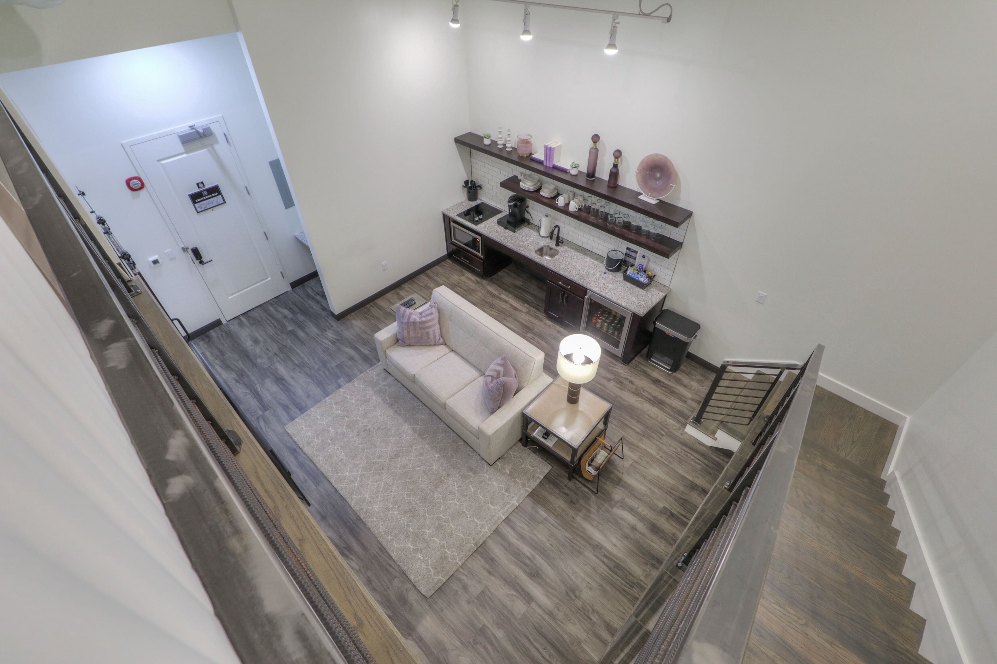 A modern kitchen with a built-in oven, a coffee machine, and a sink. There is a sofa with cushions and a small table with a lamp. A staircase is visible in the foreground.