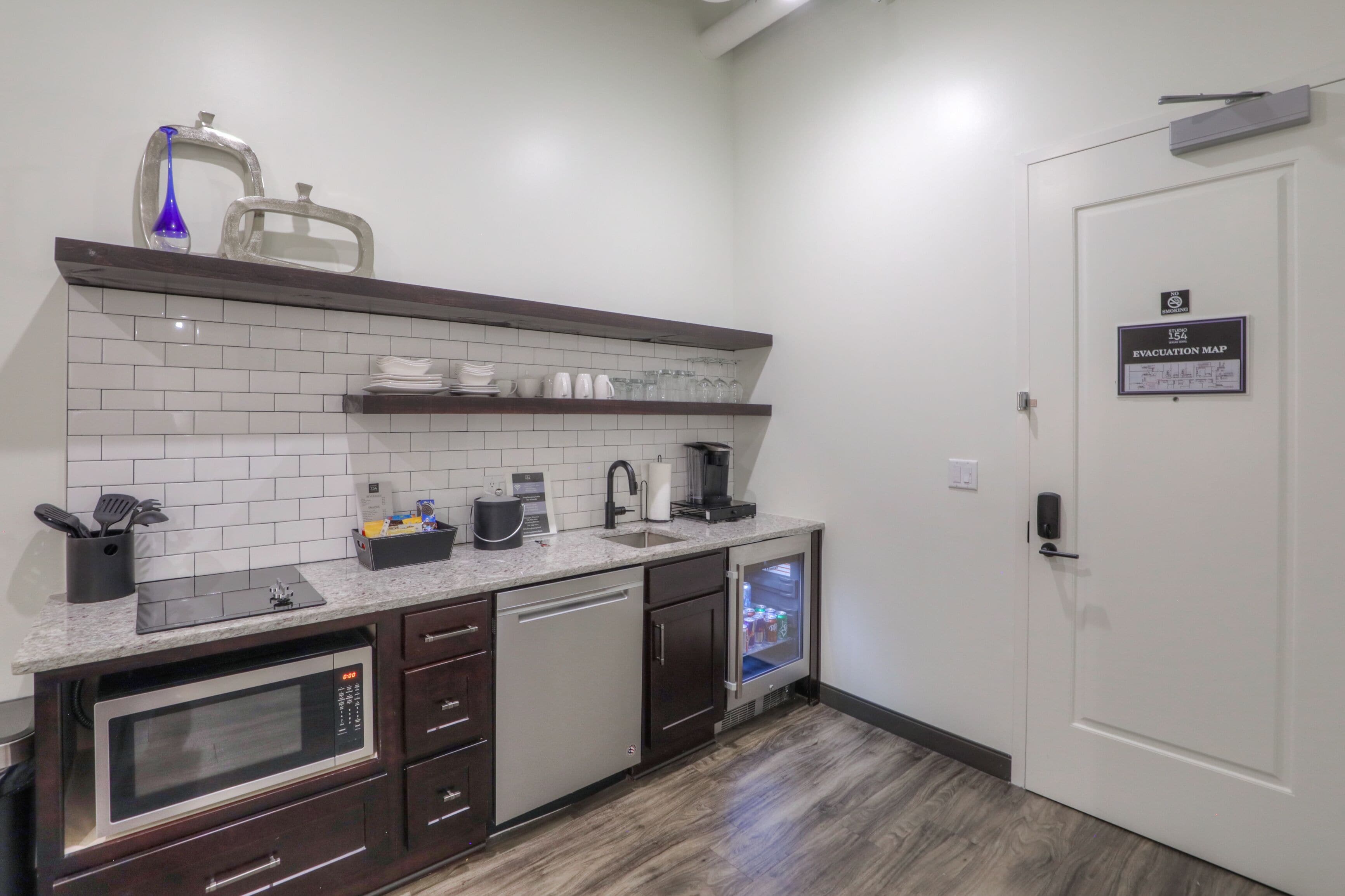 A compact kitchen with a microwave, dishwasher, and refrigerator. There is a shelf with glassware and a coffee maker. A door with an evacuation map sign is visible.