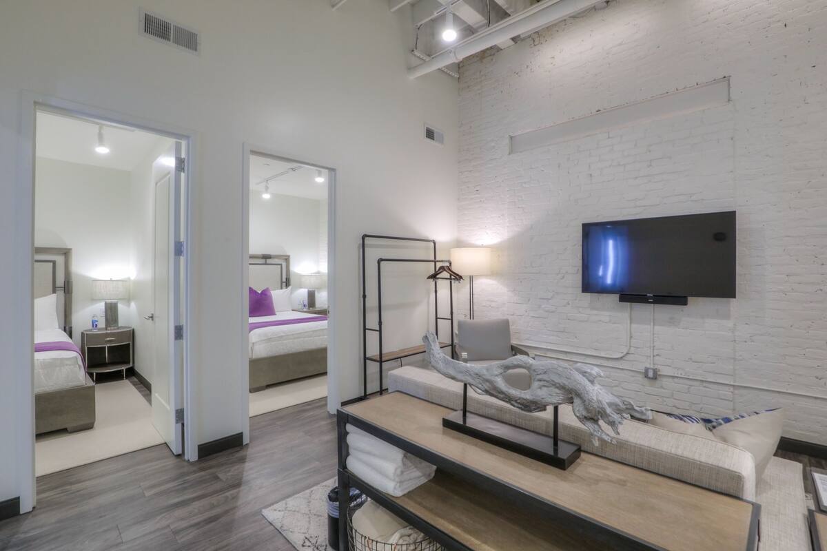 A modern hotel room with a large mirror, a flat-screen TV mounted on a brick wall, a wooden coffee table with a decorative sculpture, and a comfortable sofa.