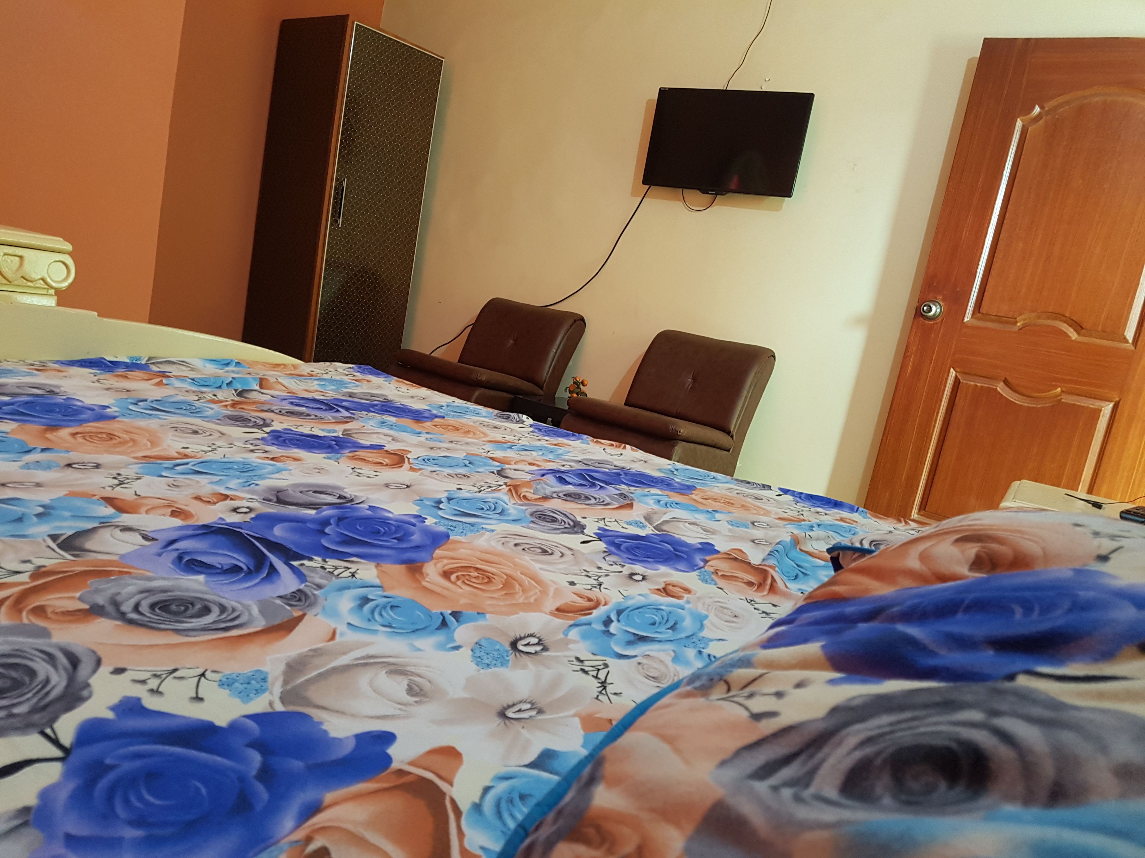 A room with a bed covered in a floral bedspread, a television mounted on the wall, and two chairs.