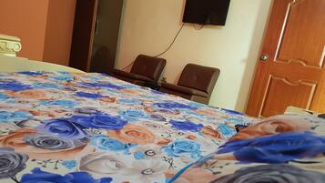 A room with a bed covered in a floral bedspread, a television mounted on the wall, and two chairs.