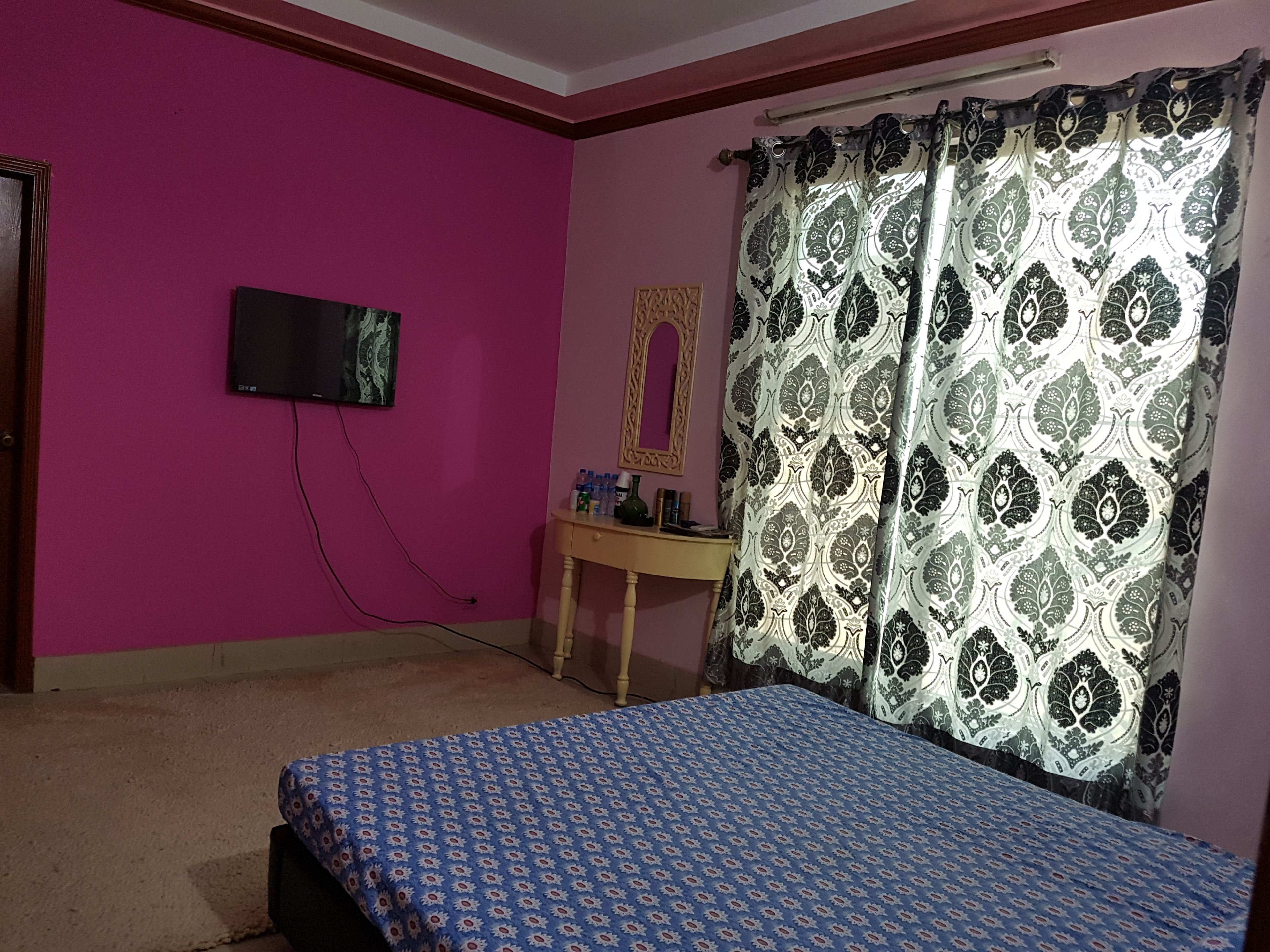 A room with a bed, a television mounted on the wall, a desk with items on it, and a patterned curtain.