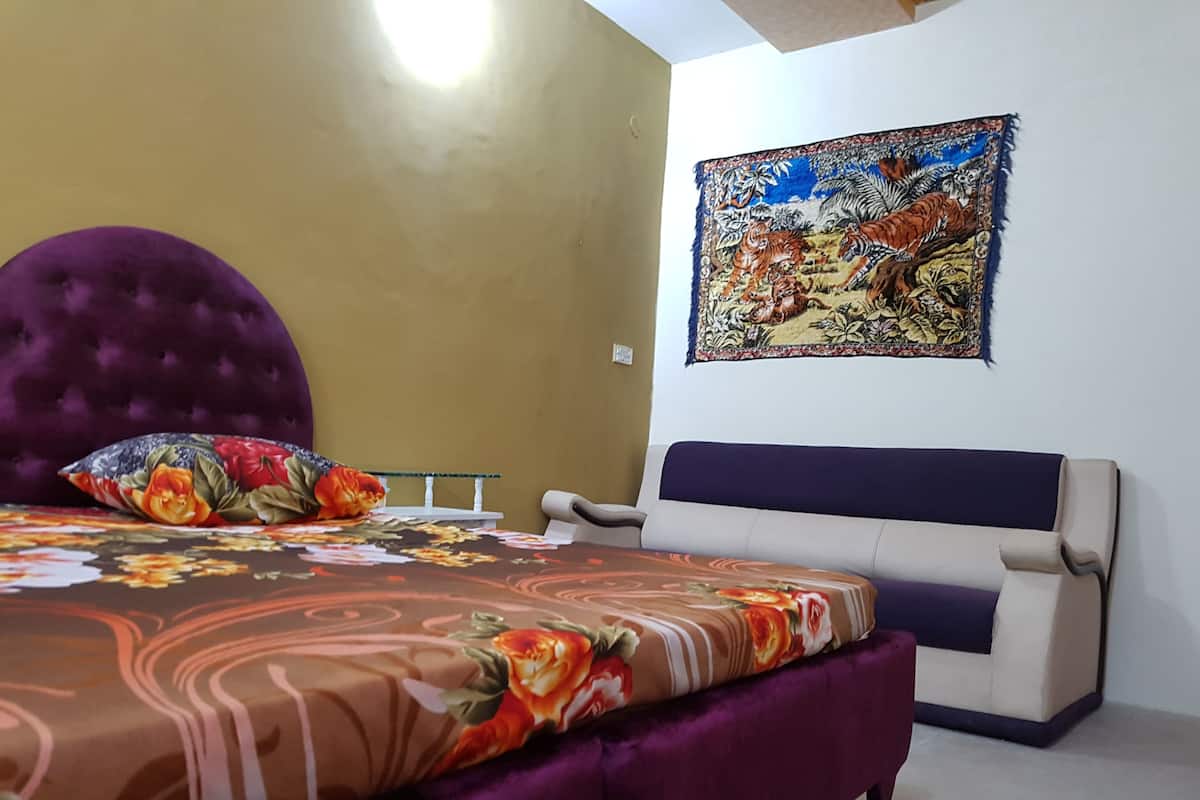 A bedroom with a bed, a purple headboard, a floral bedspread, a white armchair, and a tapestry on the wall.