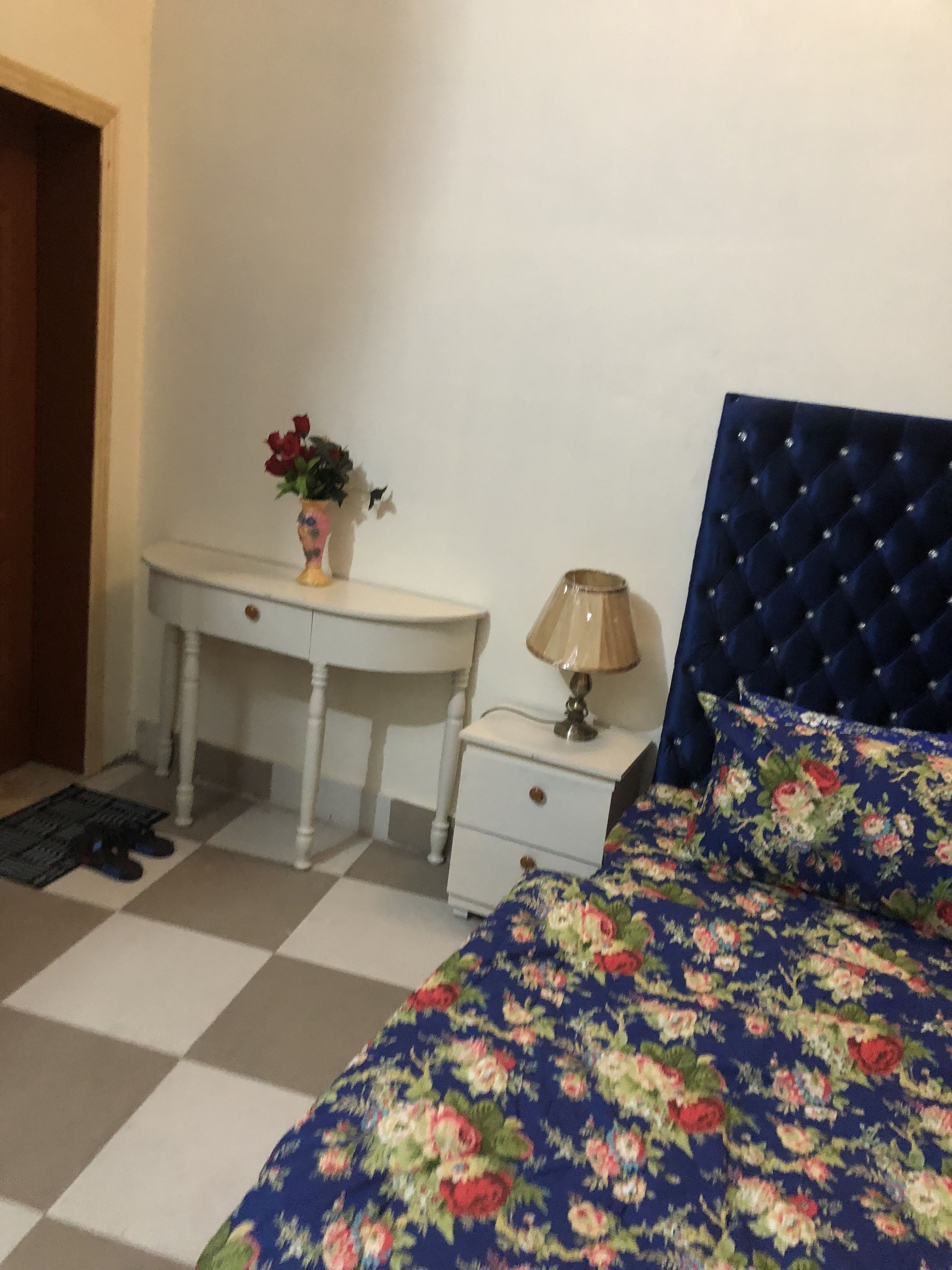 A bedroom with a checkered floor, a bed with a floral duvet, a white desk with a vase of flowers, and a nightstand with a lamp.