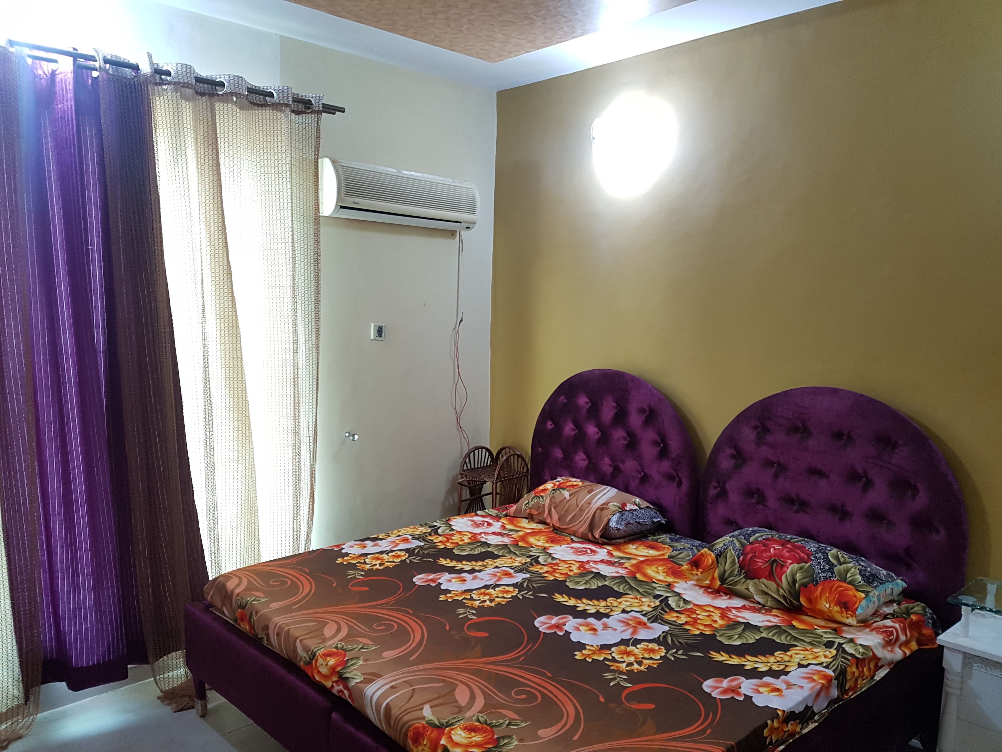 Ein Schlafzimmer mit Bett, violettem Kopfteil, floralem Bettbezug und einem Fenster mit Vorhängen.