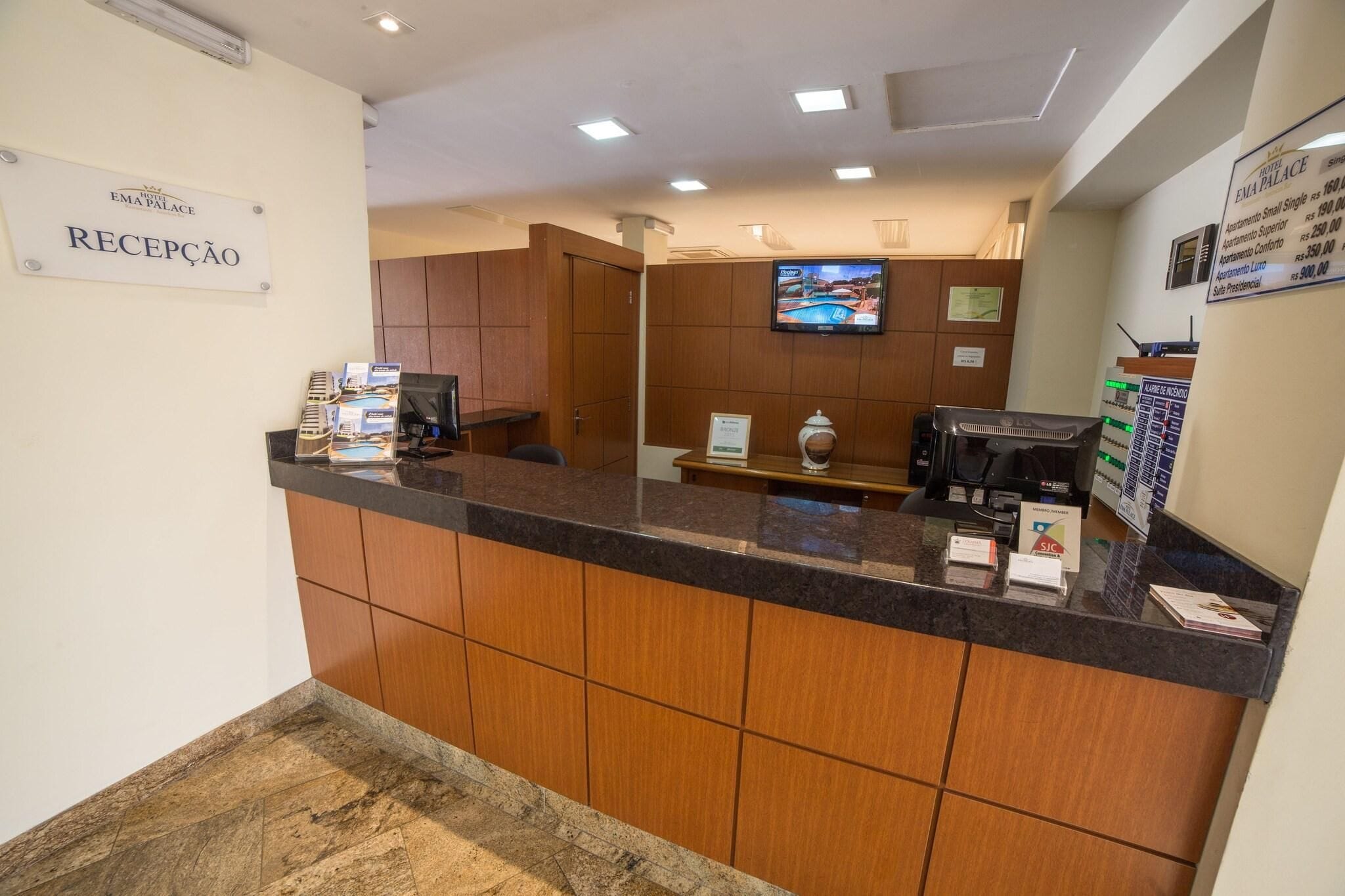 A reception area with a sign reading "RECEPCAO," a reception desk, a television mounted on the wall, and a bulletin board with property listings.