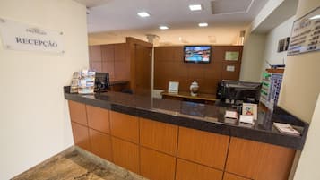 A reception area with a sign reading "RECEPCAO," a reception desk, a television mounted on the wall, and a bulletin board with property listings.