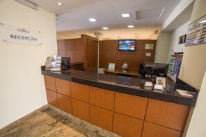 A reception area with a sign reading "RECEPCAO," a reception desk, a television mounted on the wall, and a bulletin board with property listings.