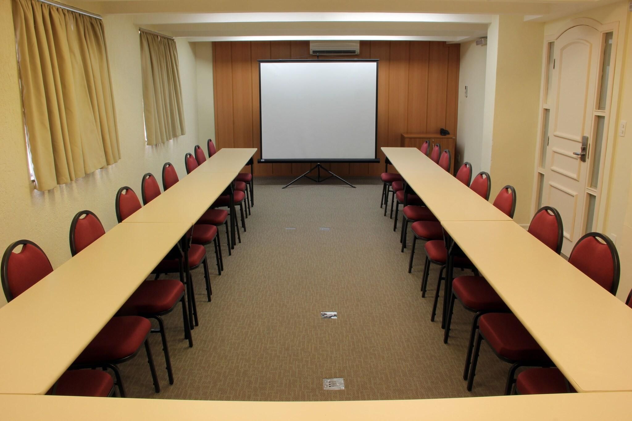 A conference room with a long table, red chairs, a whiteboard, and a projector screen.