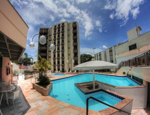 A swimming pool area with a covered seating area, a tall building in the background, and a clear sky. - Ema Palace (São José dos Campos)