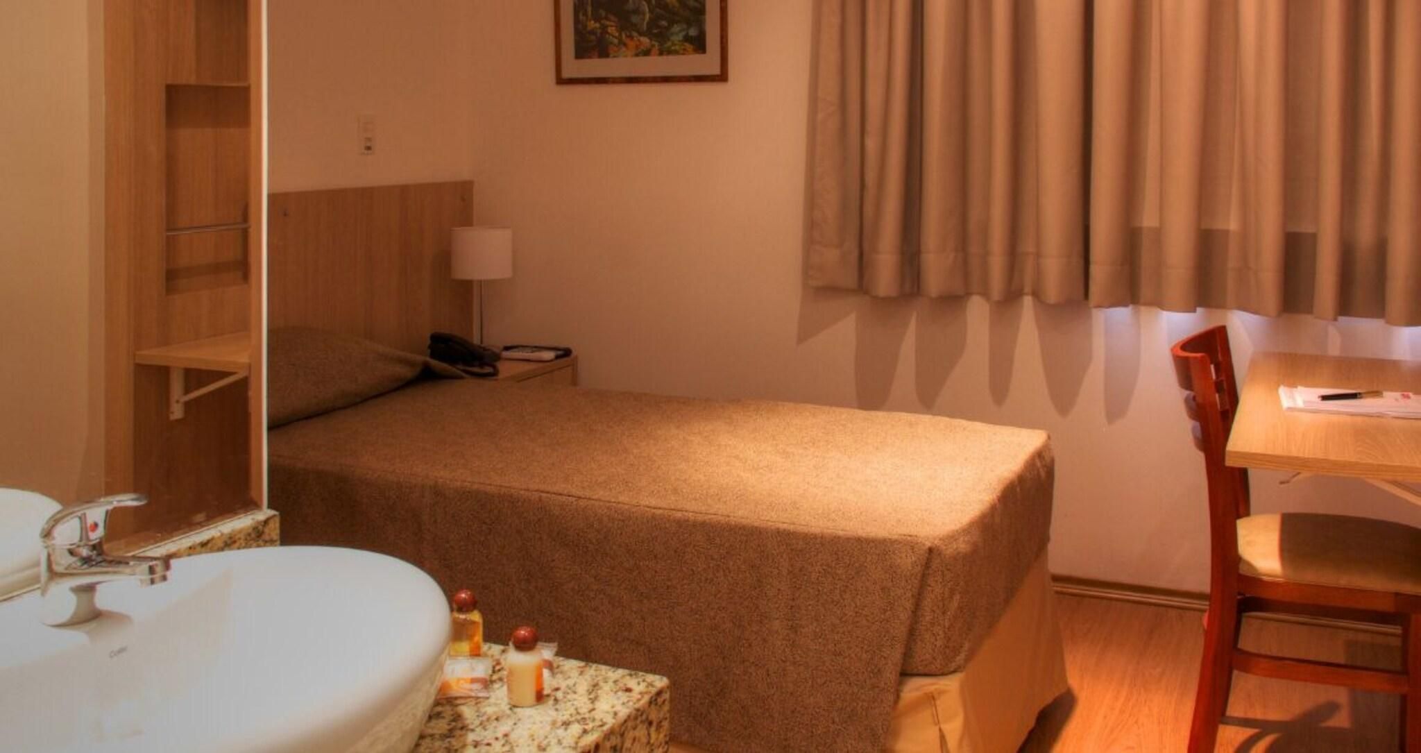 A hotel room with a bed, a desk, a chair, a bathroom with a sink, and a mirror.