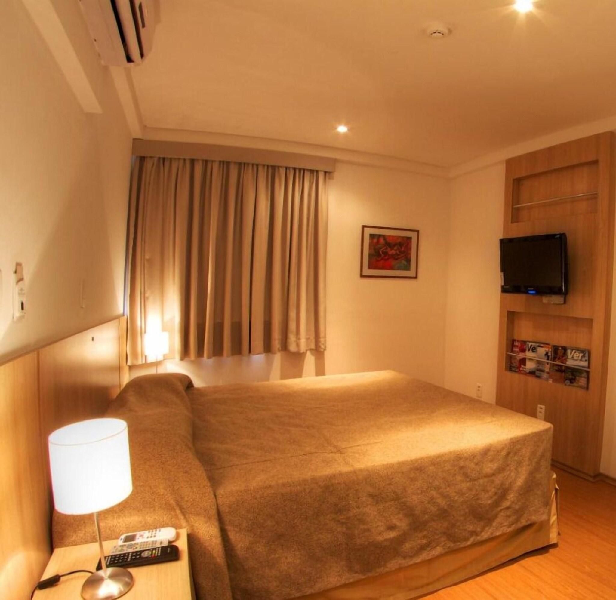 A hotel room with a bed, a TV, a lamp, and a painting on the wall.