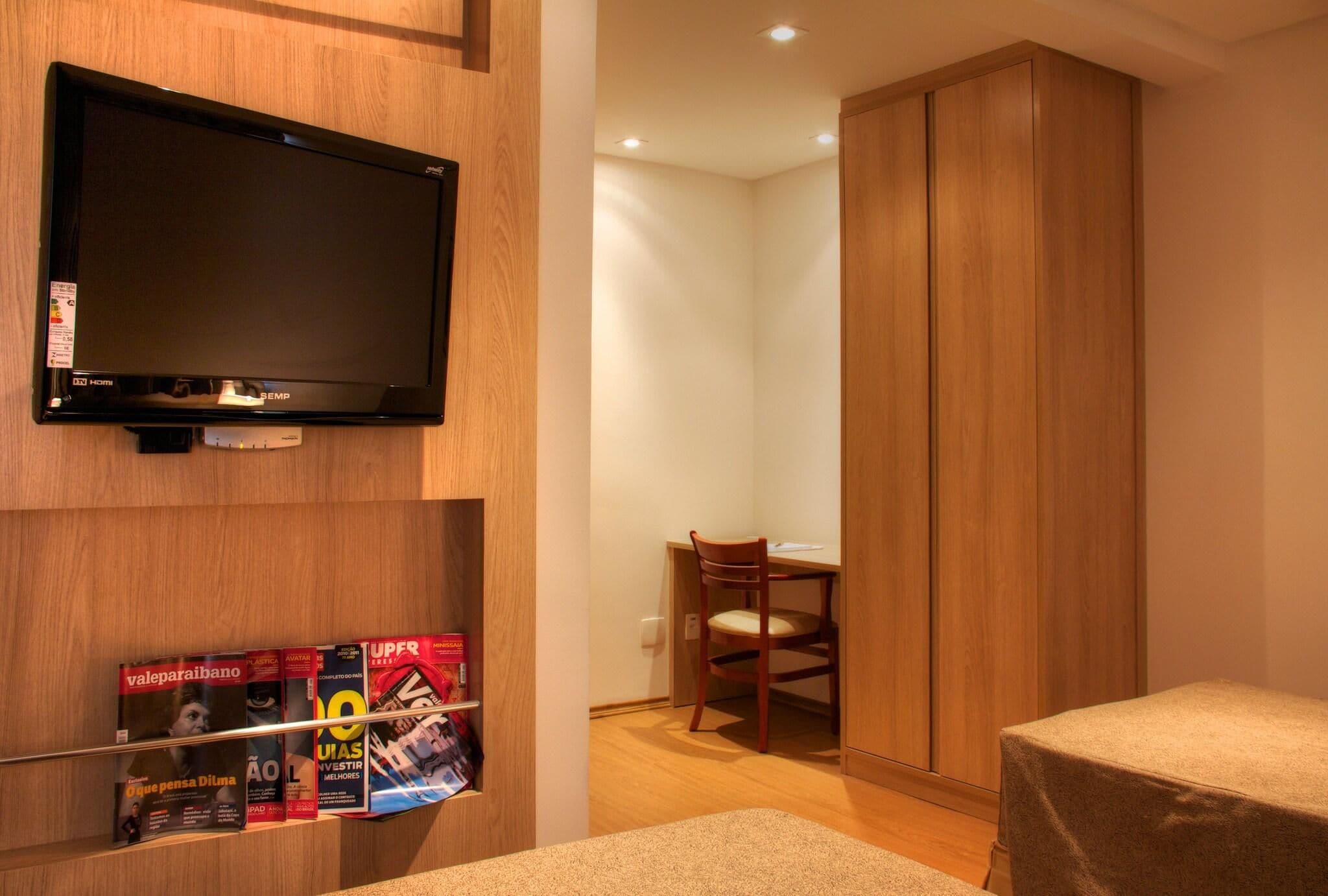 A hotel room with a television mounted on the wall, a wooden wardrobe, a desk with a chair, and a bed with a table.