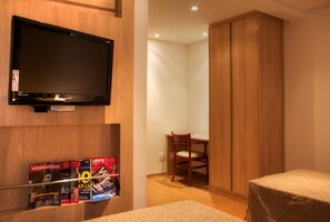 Une chambre d’hôtel équipée d’un téléviseur fixé au mur, d’une armoire en bois, d’un bureau avec une chaise et d’un lit avec une table de chevet.