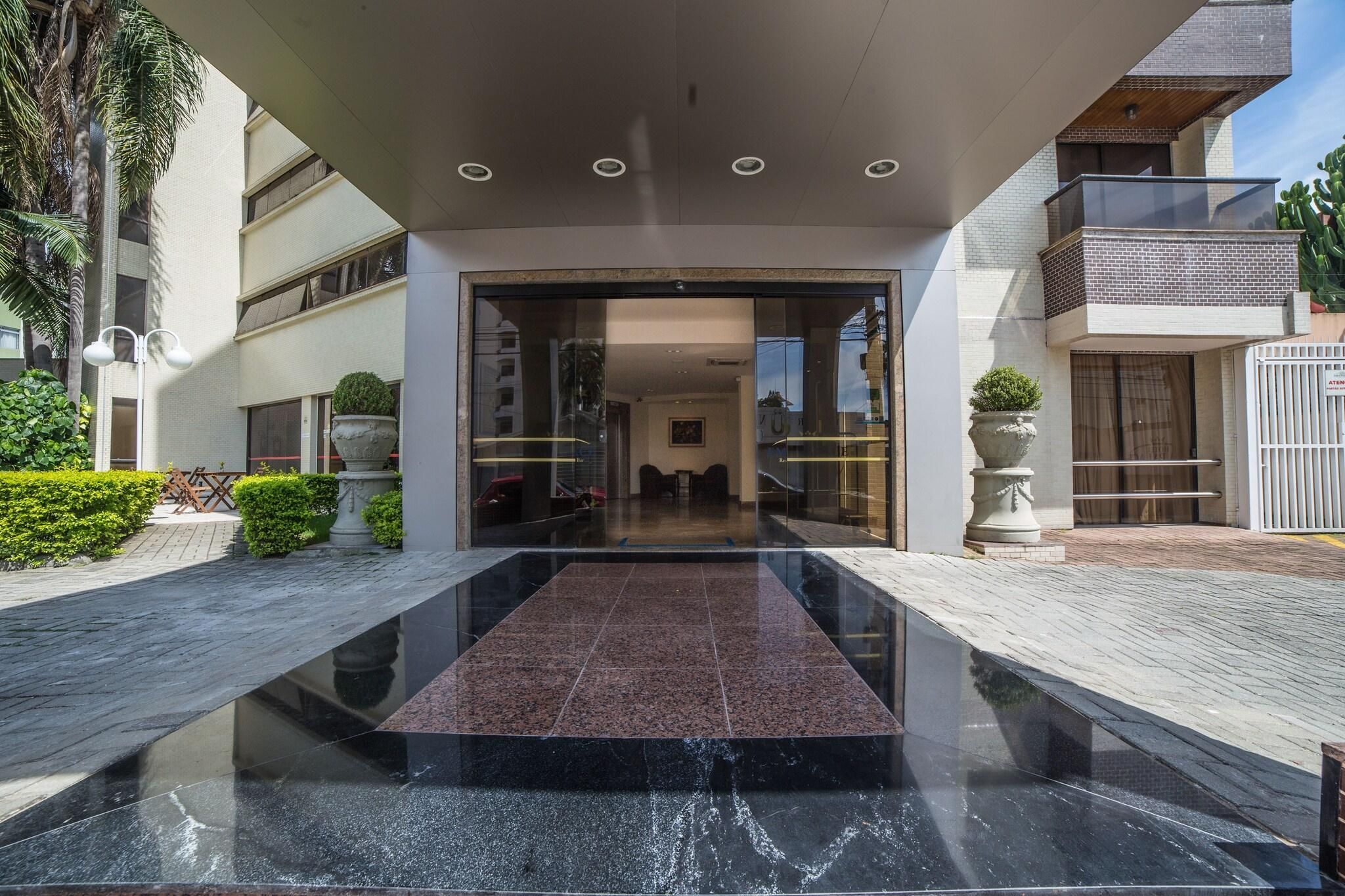 A modern hotel entrance with a glass door, a marble floor, and a small garden area with a palm tree and potted plants.