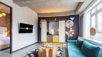 A modern hotel room with a large window, a sofa, a coffee table, and a TV displaying the hotel name.