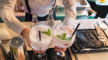Un barman sostiene dos vasos de una bebida espumosa con hojas de menta, una botella y un utensilio de bar sobre un tapete negro.