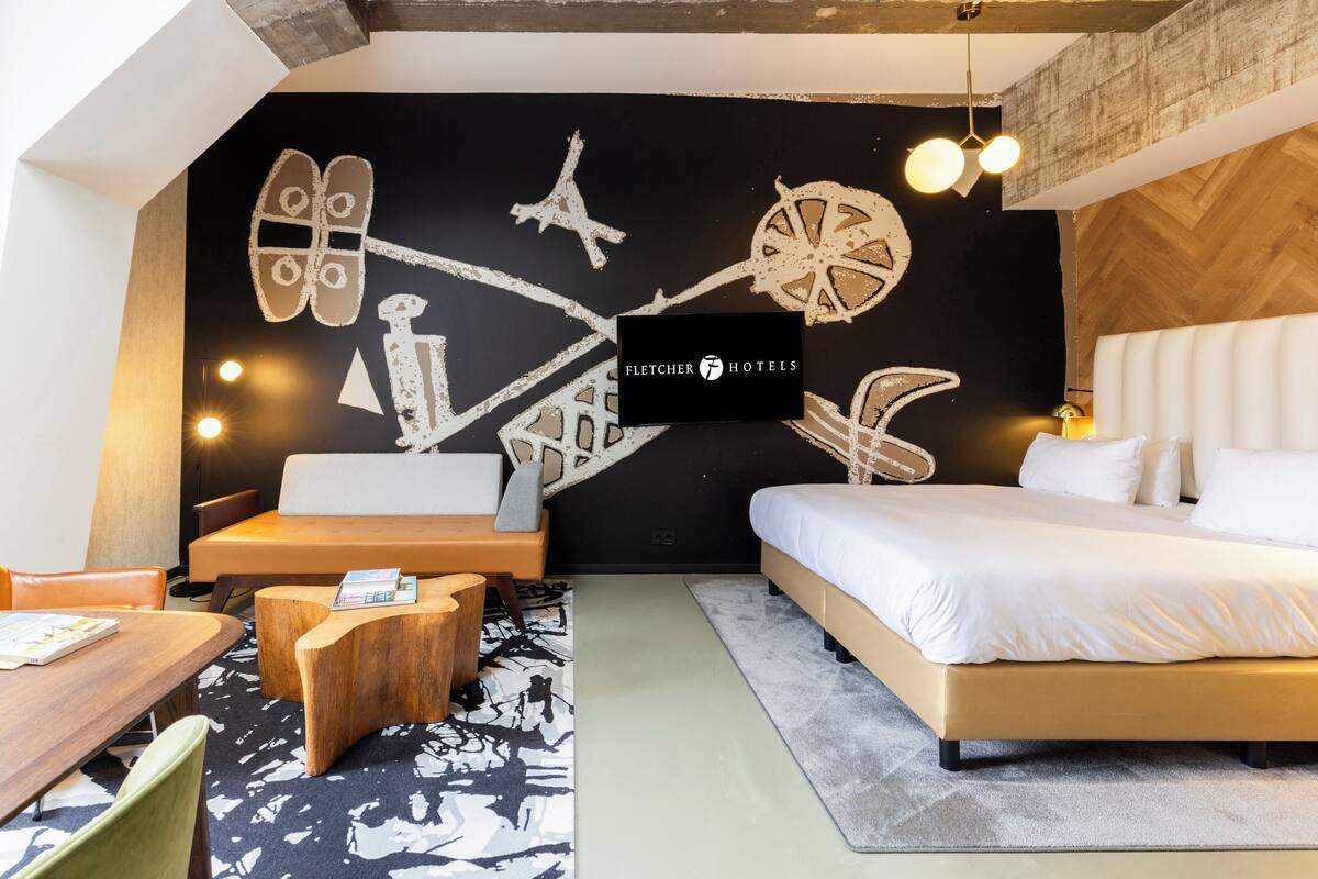 Une chambre d’hôtel moderne dotée d’un grand lit, d’un bureau et d’un coin salon. Un mur présente un décor original, et un panneau indique qu’il s’agit d’un hôtel Fletcher.